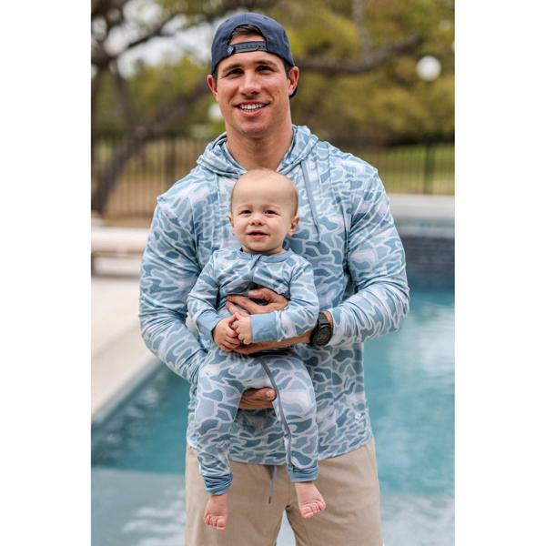 Man holding a baby both wearing matching blue camouflage outfits by a pool.