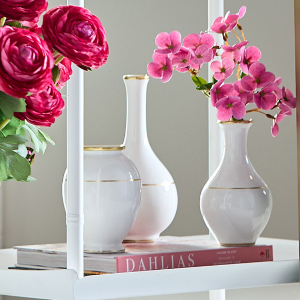 Three white vases with gold accents on a shelf with pink flowers and books.