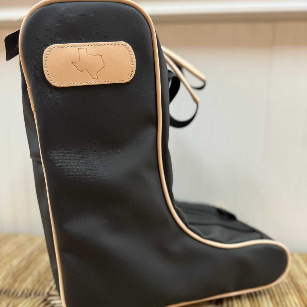 A black boot bag with natural leather trim, featuring a zippered closure and webbing handles. A tan boot is visible inside the bag, and the Texan flag is embossed on the natural leather patch on the front.