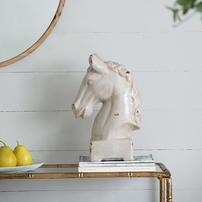 White ceramic horse head sculpture on a gold console table with a white wall and round mirror in the background.