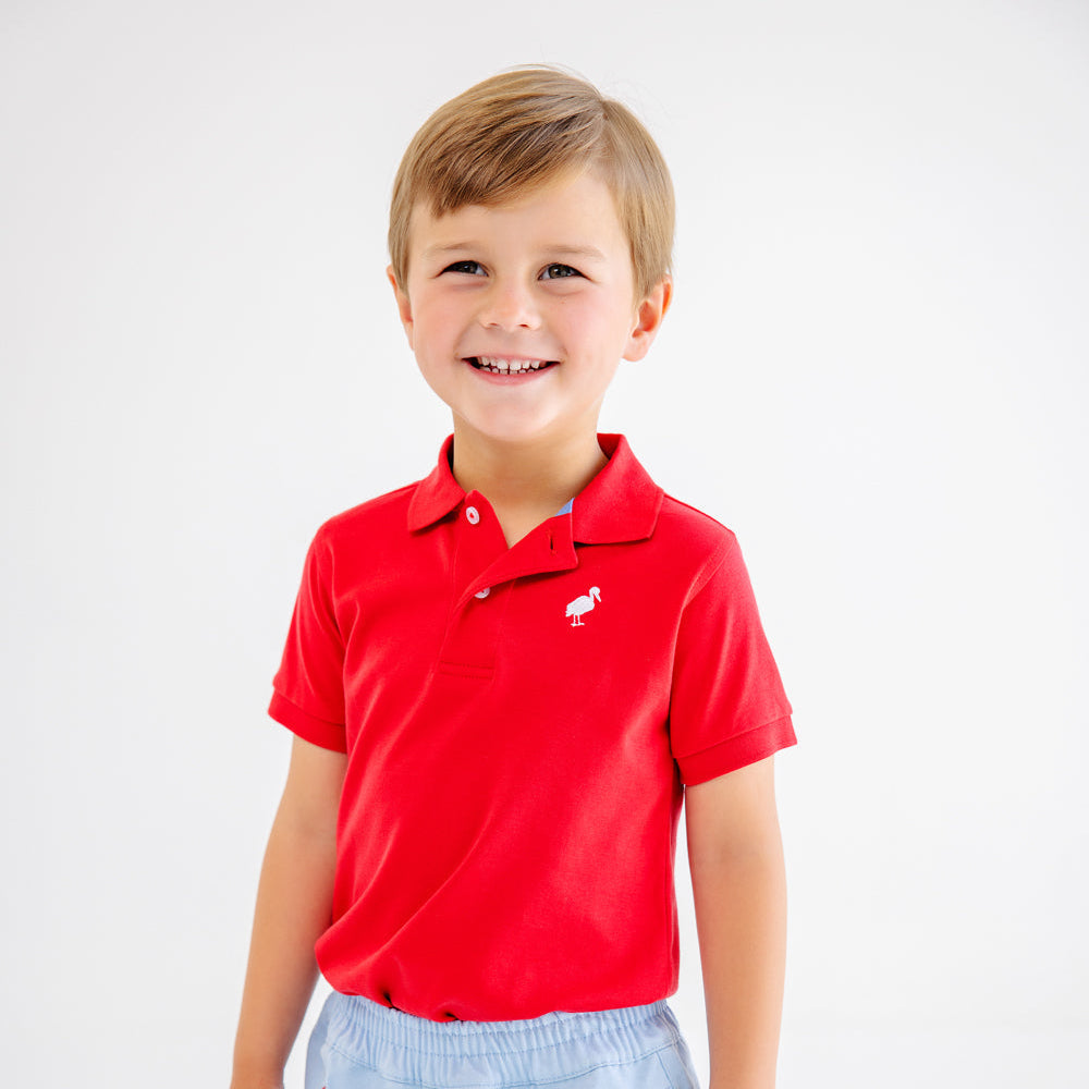 Young boy wearing a red polo shirt and light blue shorts with sailboat pattern on a white background