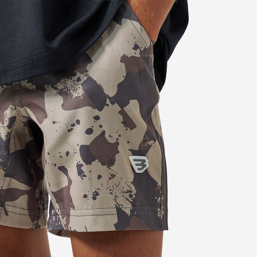 Close-up of a person wearing camouflage board shorts with a small white embroidered logo on the lower left leg, paired with a dark navy shirt against a light gray background.