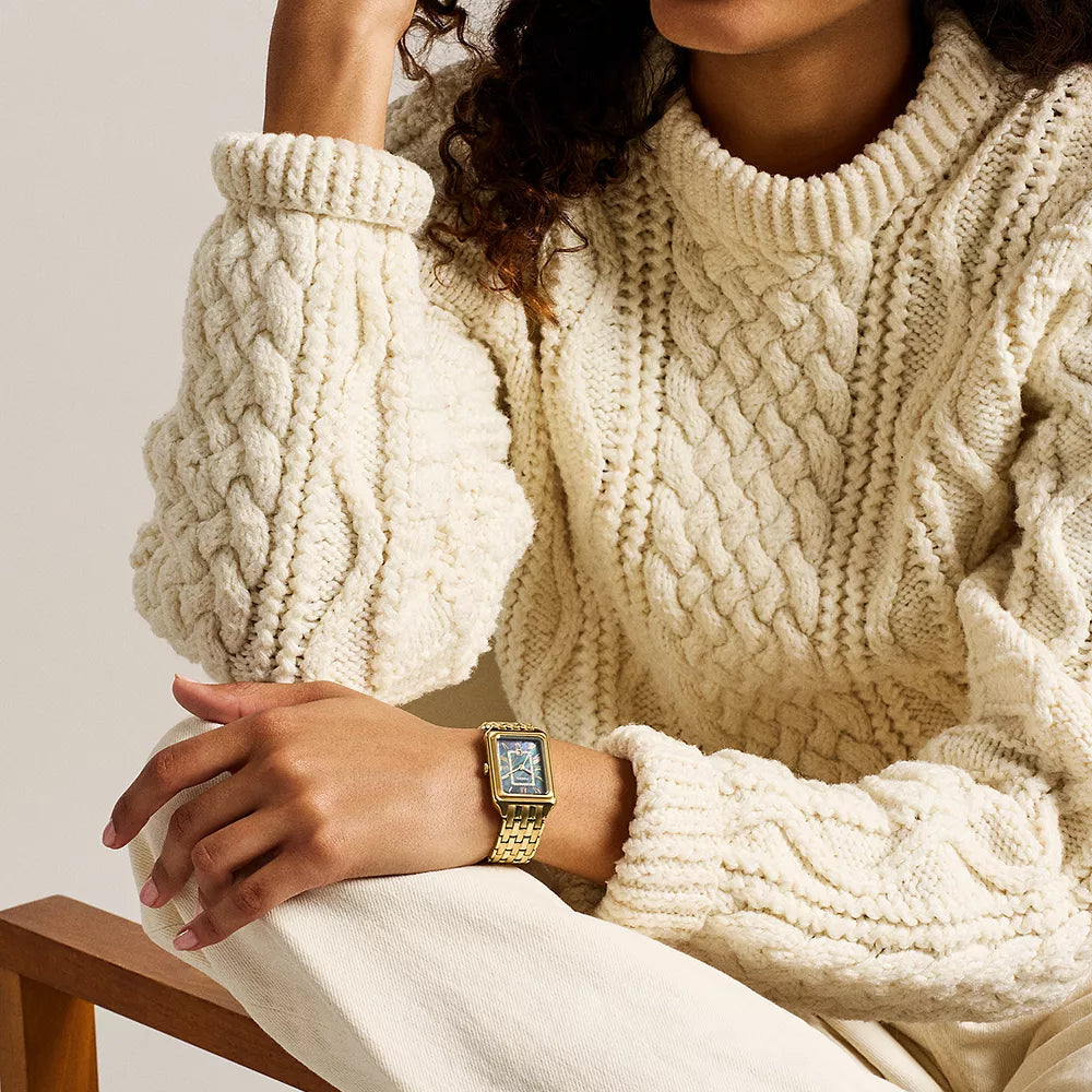 Woman wearing a cream cable knit sweater with a gold Fossil watch sitting on a wooden chair.