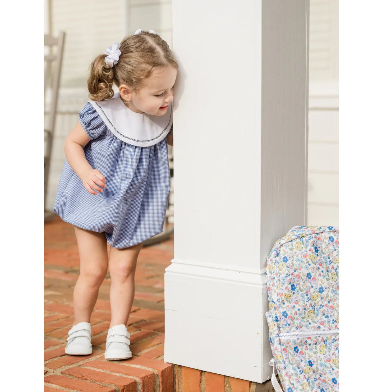 Young girl in a blue dress standing next to a white column on a brick floor.
