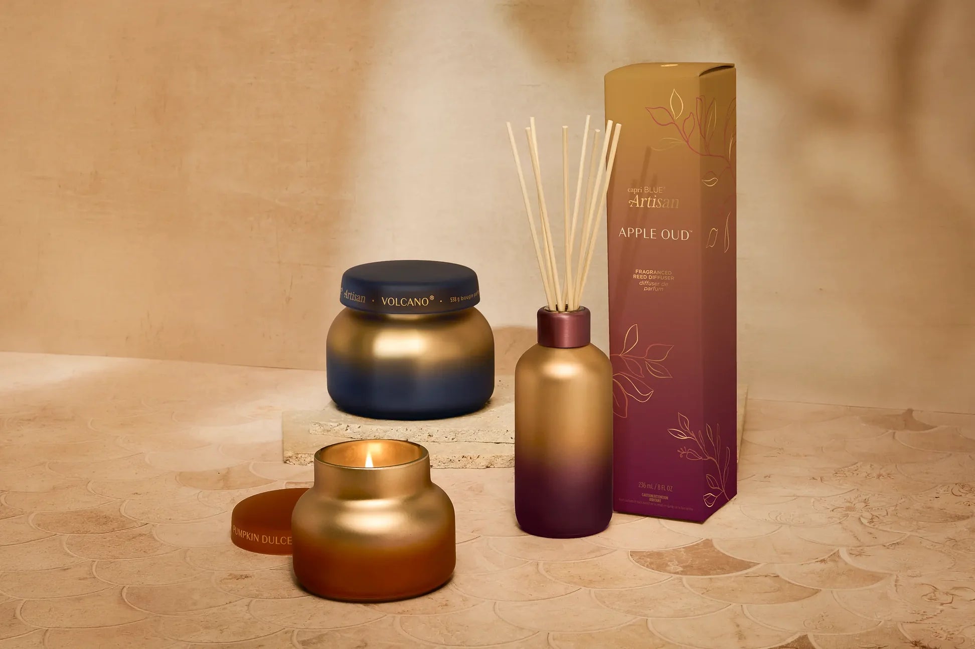 A Capri Blue reed diffuser set with a metallic amber glass bottle, matching diffuser reeds, a dark blue jar candle, and a gold-and-purple Apple Oud box, styled on a textured beige surface against a warm beige background.