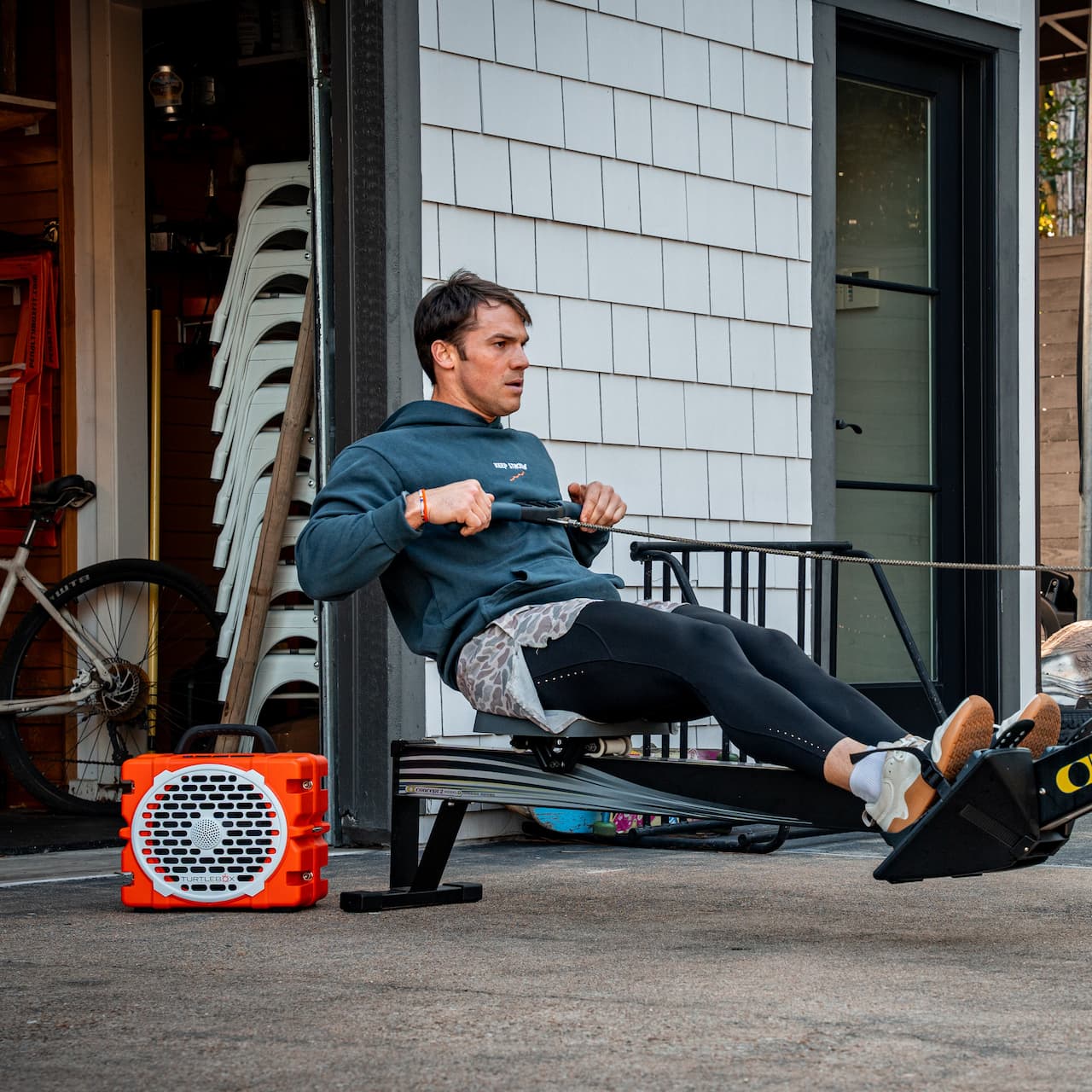 A lifestyle image from a man using a rowing machine outdoors next to an orange Grande with a building and trees in the background #speaker-color_orange