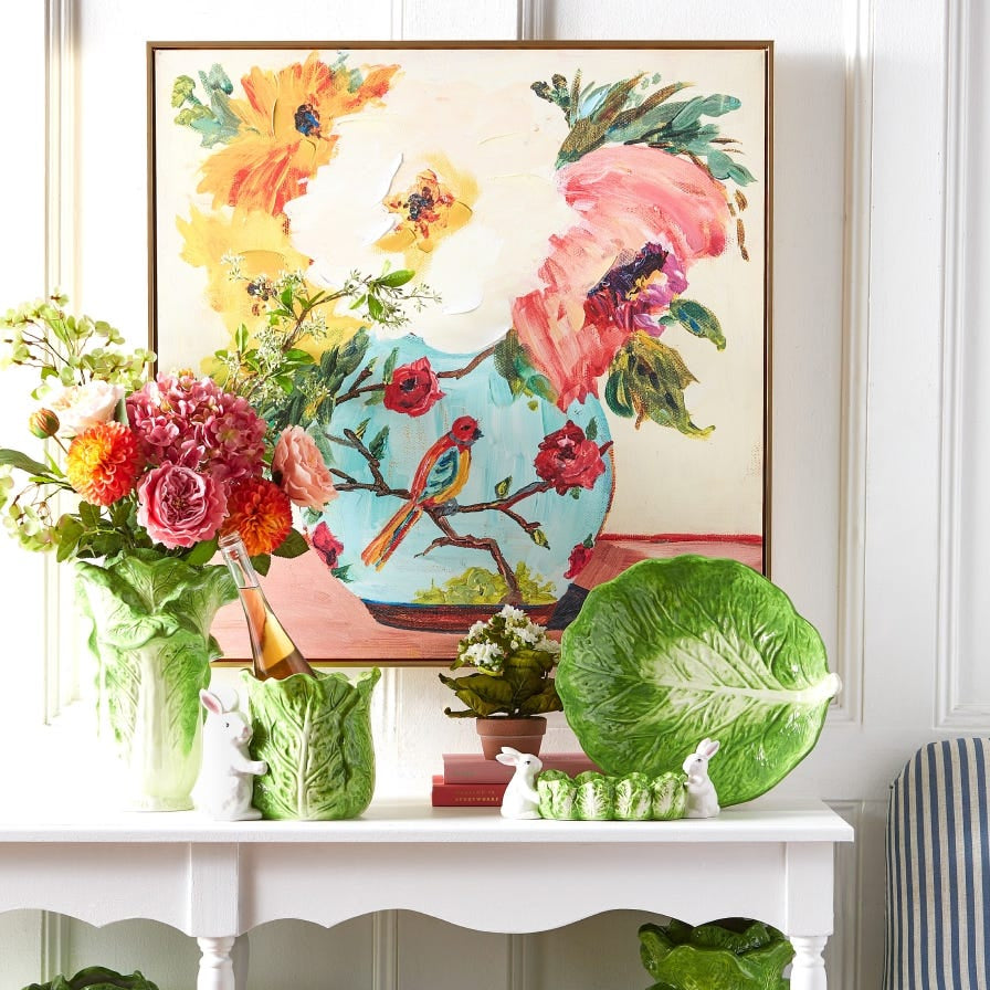 Decorative table with floral arrangements, a large green leaf, and a colorful painting of flowers and birds on a white wall.
